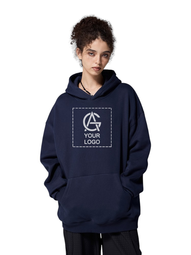 A person with curly hair wears a loose-fitting navy hoodie featuring a placeholder square with “YOUR LOGO” and a stylized logo in the center. The person stands with hands in the hoodie’s pockets against a white background.