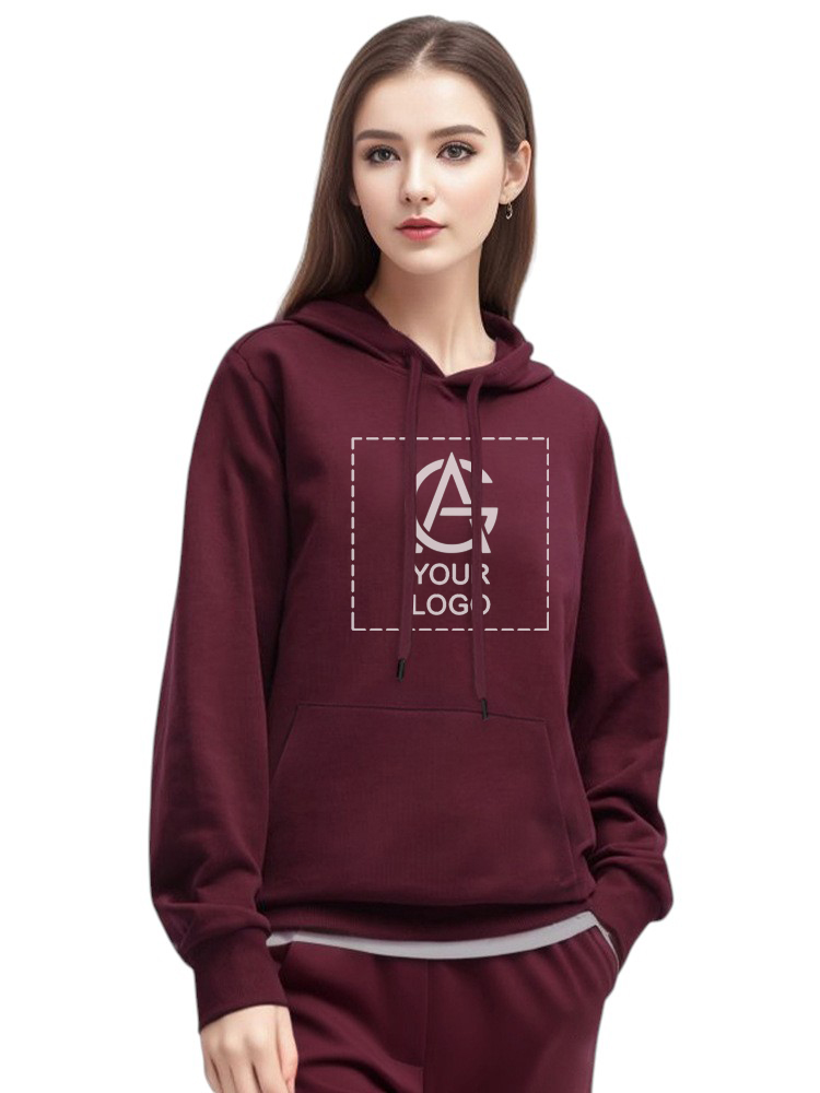 A woman wearing a maroon hoodie with a customizable logo area on the front stands facing forward. The hoodie has a front pocket and drawstrings, and she is also wearing matching pants.