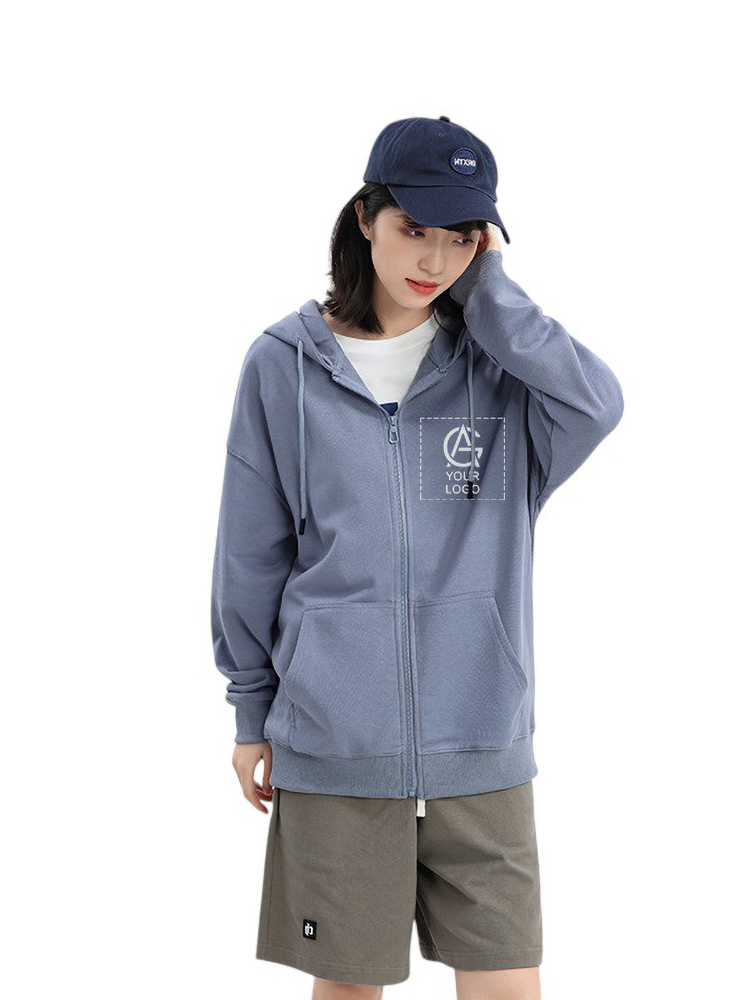A person wearing a blue zip-up hoodie, khaki shorts, and a navy blue cap stands against a plain white background, looking down with one hand touching the side of their head.