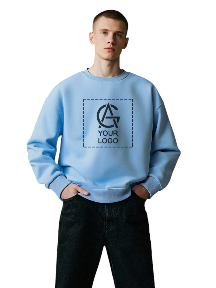 A young man poses wearing the Sweatshirt Y04, a light blue sweatshirt with a placeholder graphic and the text "YOUR LOGO" centered on the chest, paired with black pants, against a plain white background.