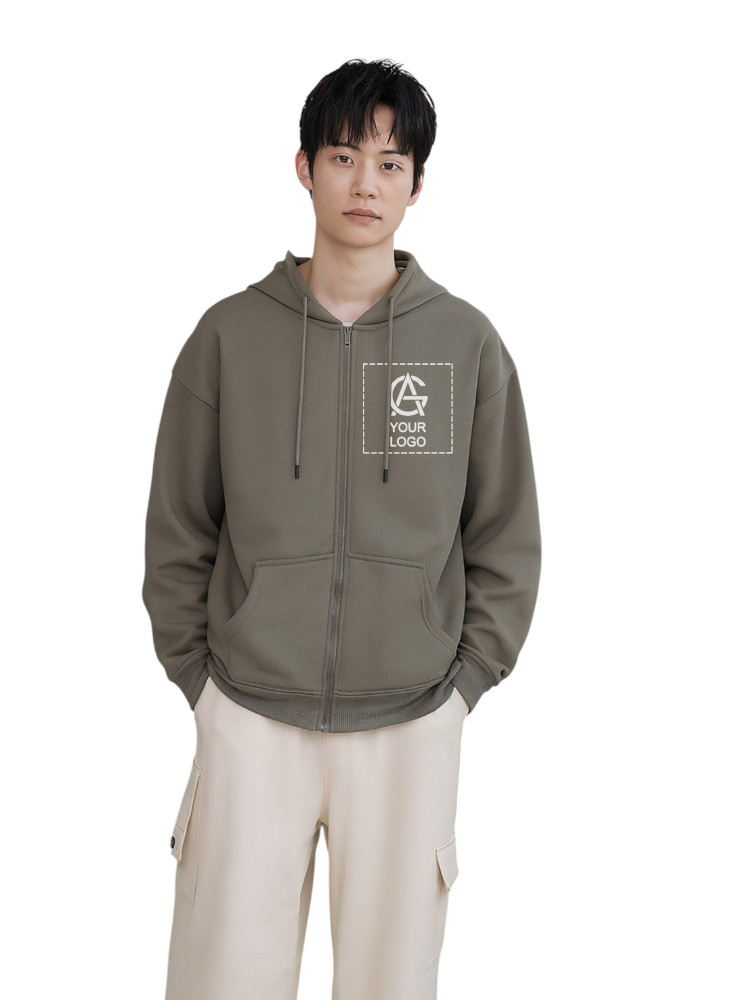 A young person with short dark hair wearing an olive green zip-up hoodie with a logo on the chest and light beige pants, standing with hands in pockets against a plain white background.