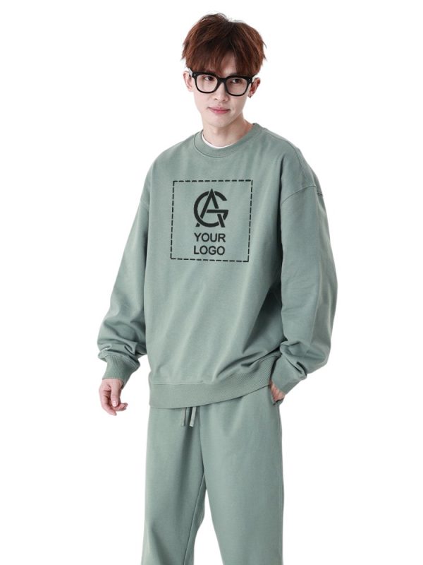 A person with short brown hair and glasses wears a loose, sage green sweatshirt and matching pants. The sweatshirt features a placeholder logo and the text "YOUR LOGO" on the front.