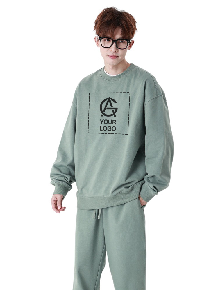 A person with short brown hair and glasses wears a loose, sage green sweatshirt and matching pants. The sweatshirt features a placeholder logo and the text "YOUR LOGO" on the front.