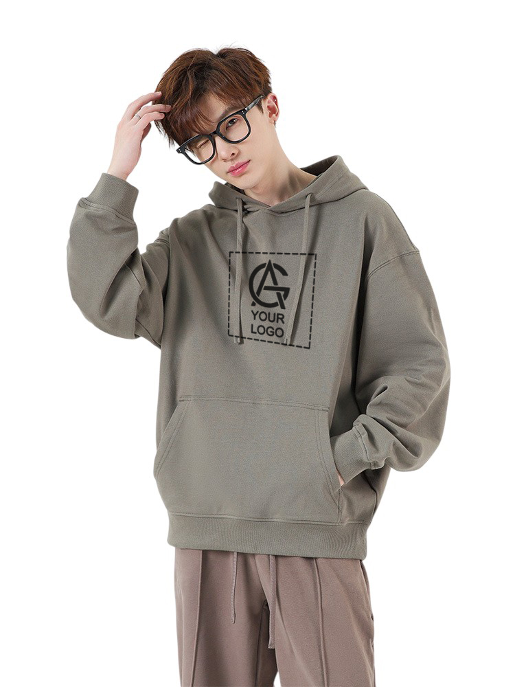 A young person with short brown hair and glasses wears a loose-fitting, olive green hoodie with a custom logo and "YOUR LOGO" text on the chest, standing with one hand on their head against a plain background.