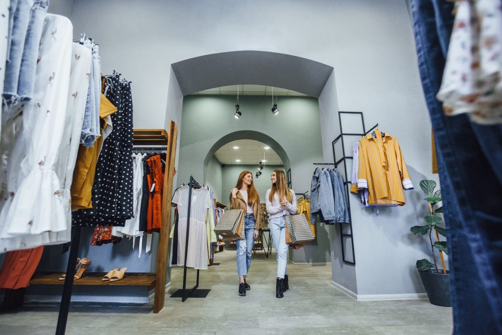 Two women holding shopping bags walk and smile together inside a stylish clothing store, surrounded by racks of colorful clothes, bright interior lighting, and displays highlighting the latest hoodie price trends.